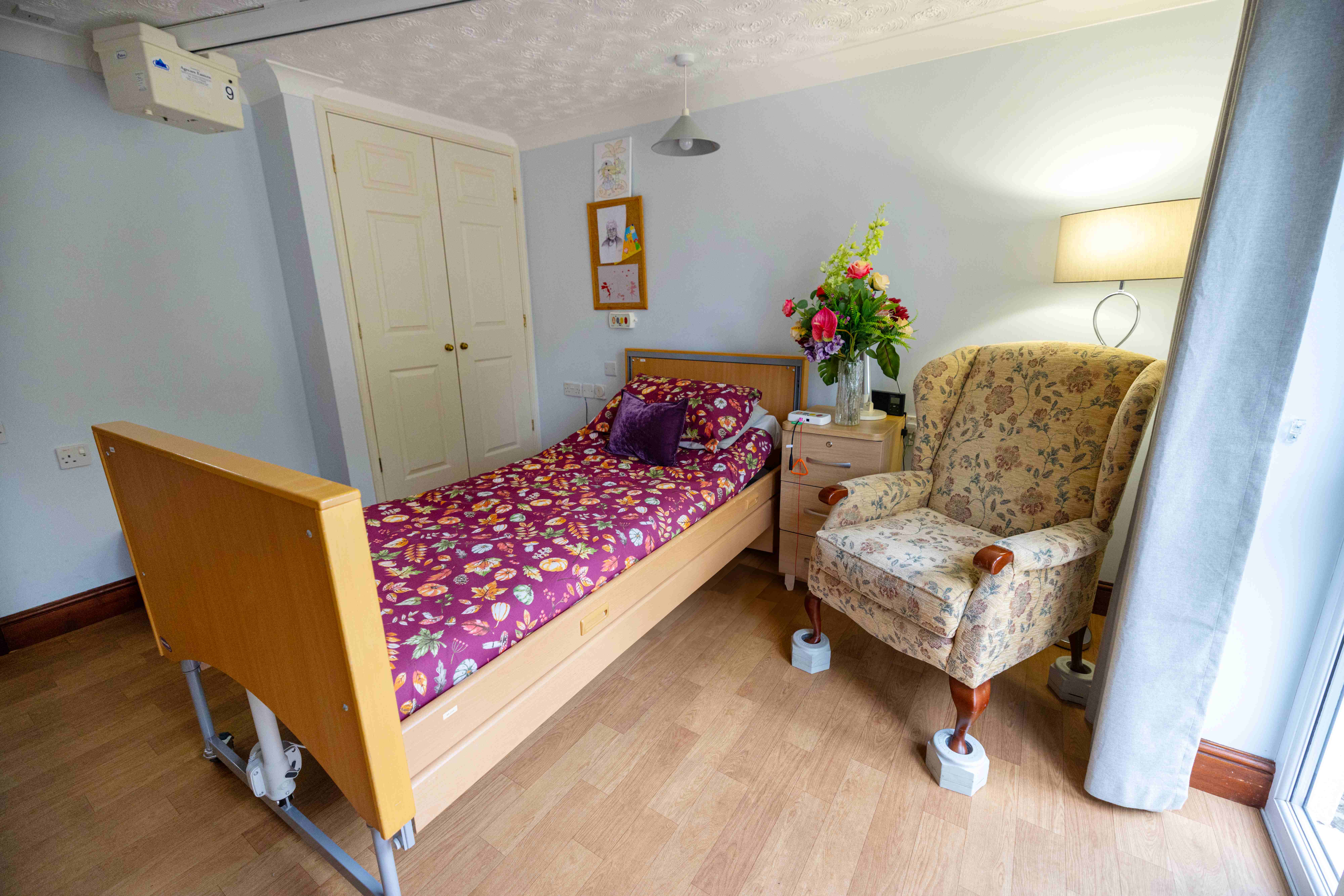 Comfortable private resident bedroom with adjustable bed, built-in wardrobe, comfortable wingback armchair, fresh flowers, and warm lighting